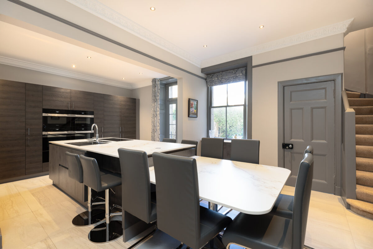 Modern open-plan kitchen and dining area with Dekton countertop island, built-in appliances, dark wood cabinets, and grey leather chairs.